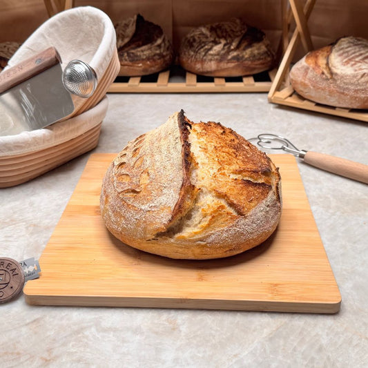 Loaf of bread on a wooden cutting board with bread-making tools in the background.